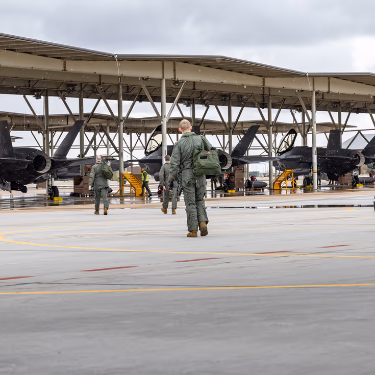 Vliegers lopen met tassen richting F-35’s die onder een overkapping op een platform staan.