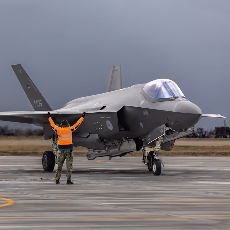 Twee militairen in oranje hesjes naast een F-35 op het platform. Een van de militairen houdt de armen schuin naar boven gespreid in de lucht.