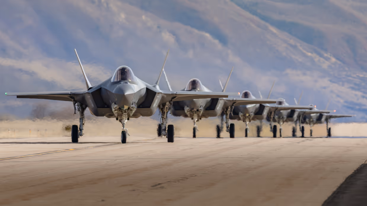 Vijf F-35’s taxiën achter elkaar op een startbaan met op de achtergrond bergen.