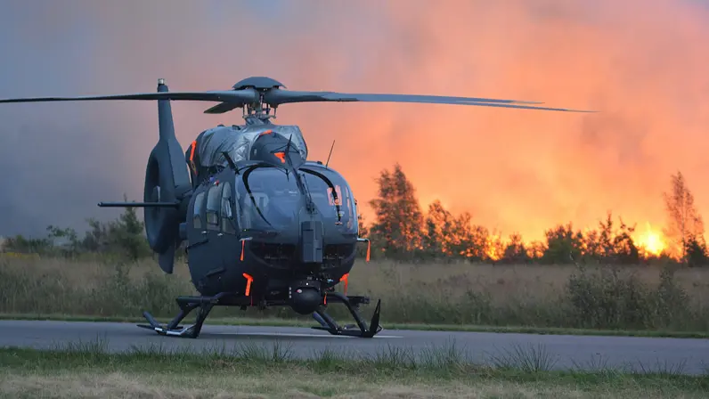 Een helikopter staat op een betonnen baan. Op de achtergrond staat het bos in de brand.    