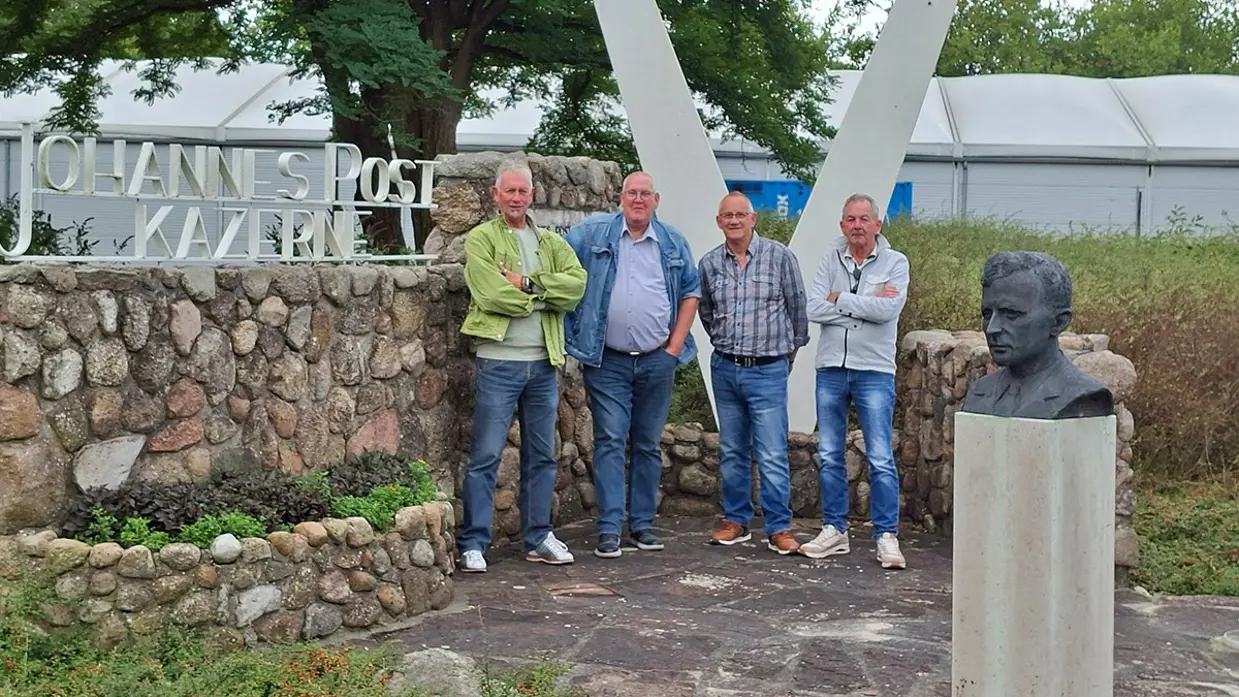 Vier oudere mannen staan met de armen over elkaar  bij een muur en daarop de woorden Johannes Postkazerne. Op de voorgrond staat een borstbeeld.