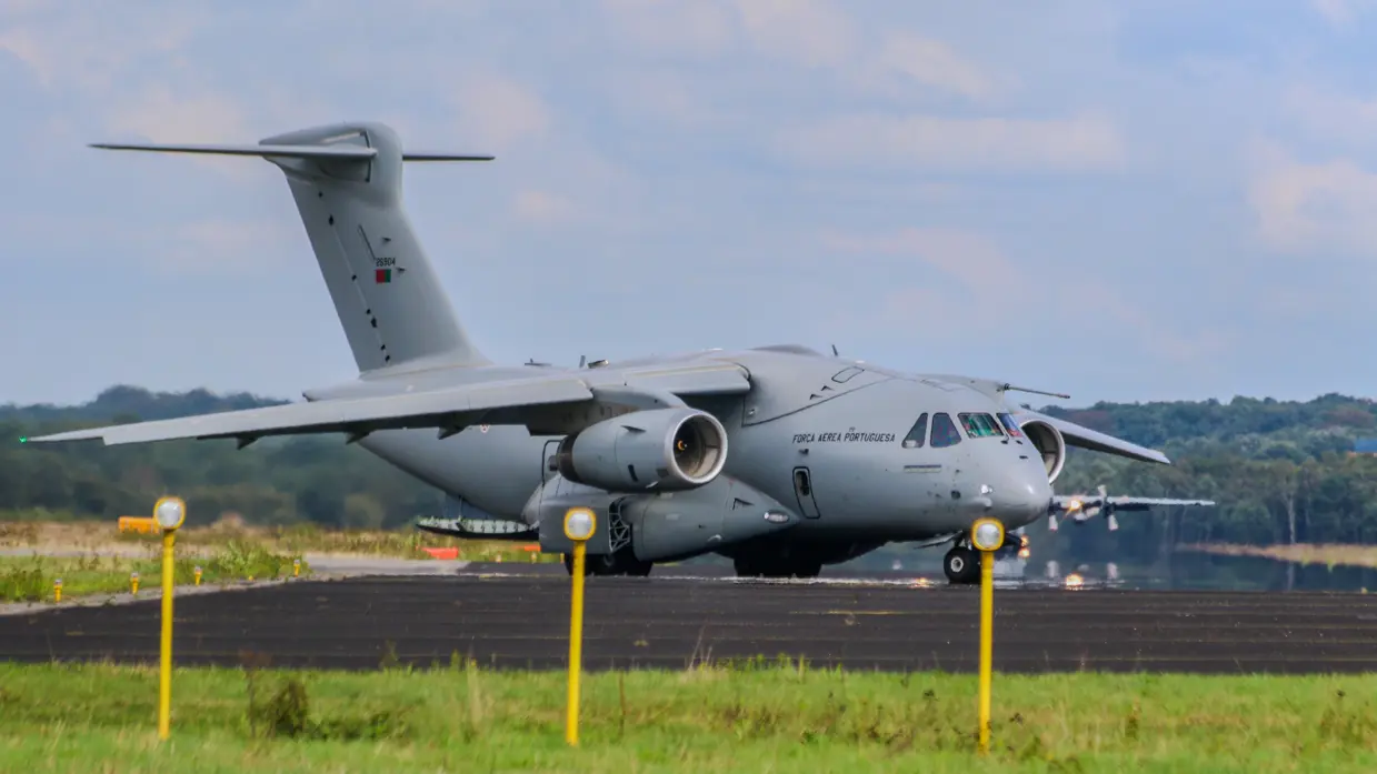 Een grijs militair transportvliegtuig van de Portugese luchtmacht staat op een startbaan met groen gras, bomen en een blauwe lucht. 