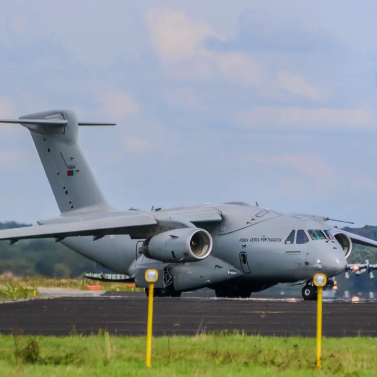 Een grijs militair transportvliegtuig van de Portugese luchtmacht staat op een startbaan met groen gras, bomen en een blauwe lucht. 