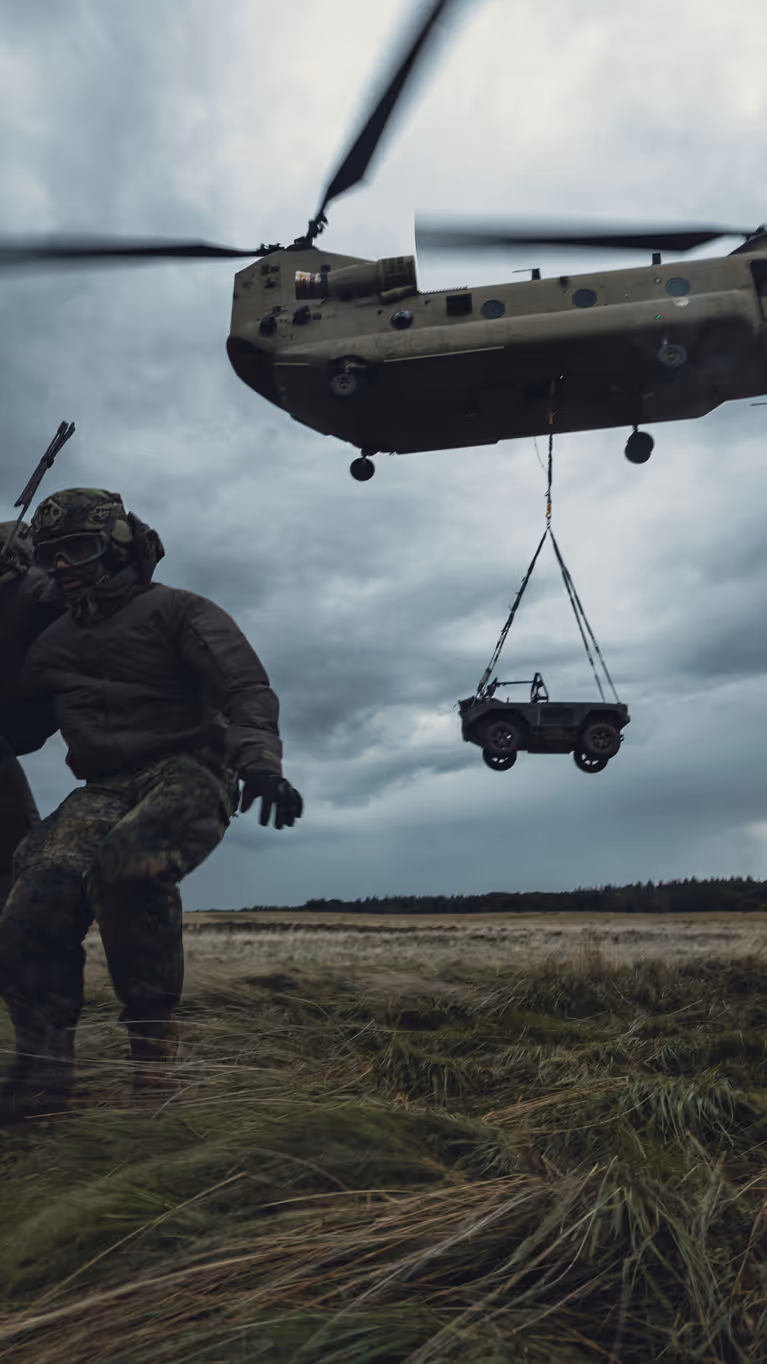 Een Chinook-transporthelikopter stijgt op met een voertuig die eronder hangt. Drie militairen staan op de heide en houden elkaar vast om te blijven staan in het vlieggeweld.