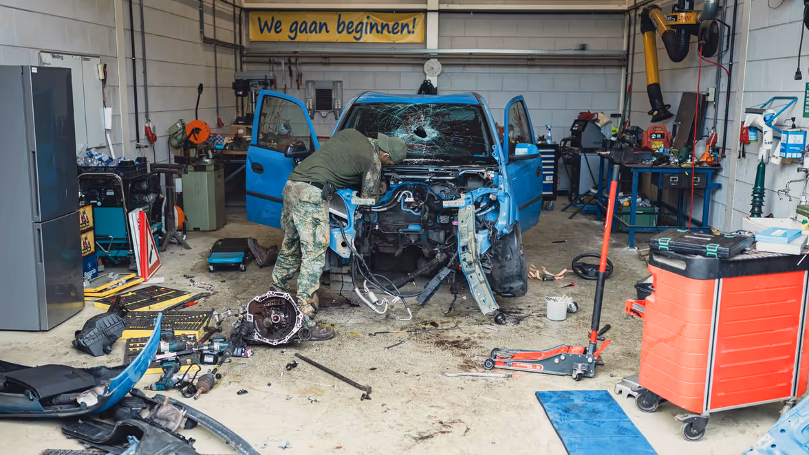 Een militair sleutelt aan een blauw voertuig. Er zit een gat in de voorruit en de auto is volledig beschadigd.