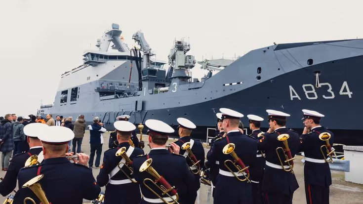 Marineschip ligt langs kade. Op voorgrond militaire muziekkapel.