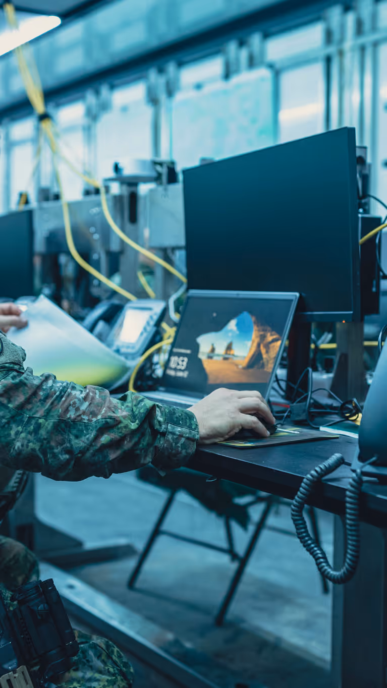 Een bewapende militair zit op een klapstoel achter een computer en bladert door een mapje.