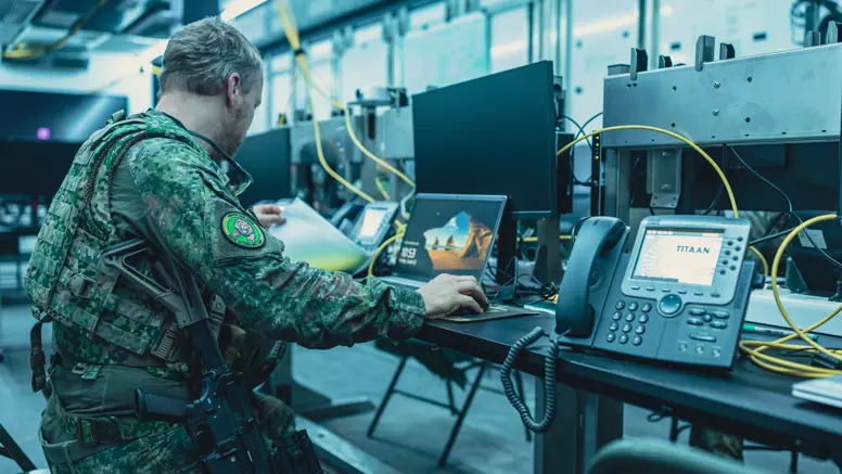 Een bewapende militair zit op een klapstoel achter een computer en bladert door een mapje.