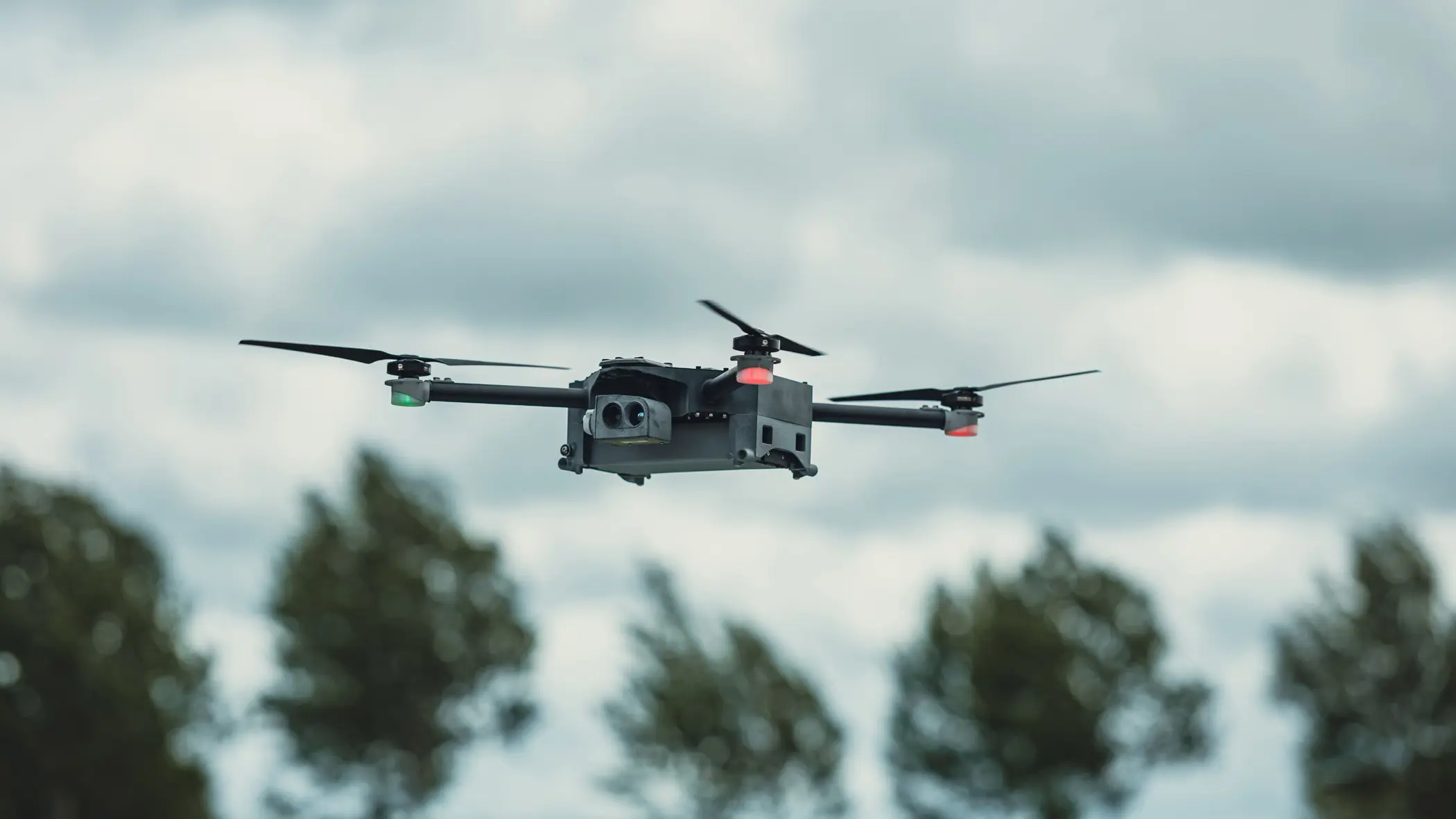 Een grijze, vliegende drone. Op de achtergrond een rij bomen.