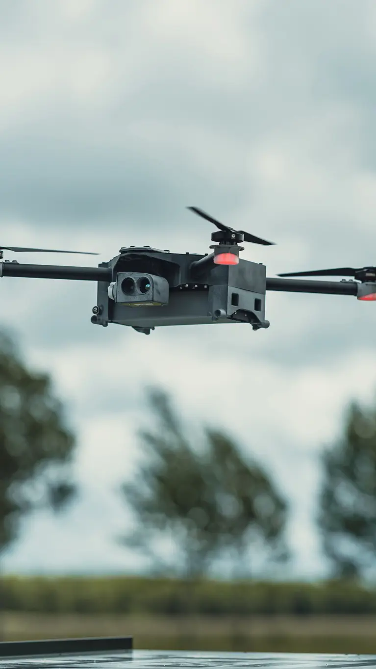 Een grijze, vliegende drone. Op de achtergrond een rij bomen.