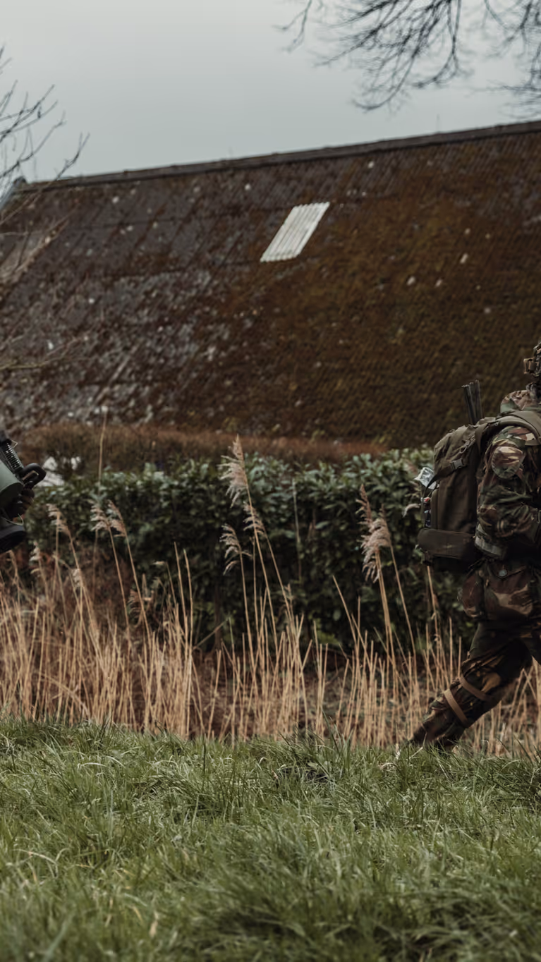 Twee gewapende militairen lopen achter elkaar door een grasveld. De achterste draagt een cilindervormig, donkergroen antitankwapen op zijn schouder.