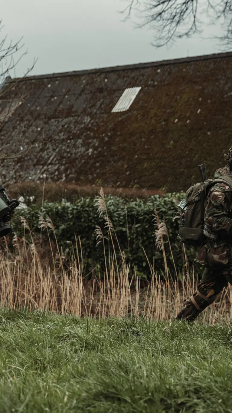 Twee gewapende militairen lopen achter elkaar door een grasveld. De achterste draagt een cilindervormig, donkergroen antitankwapen op zijn schouder.