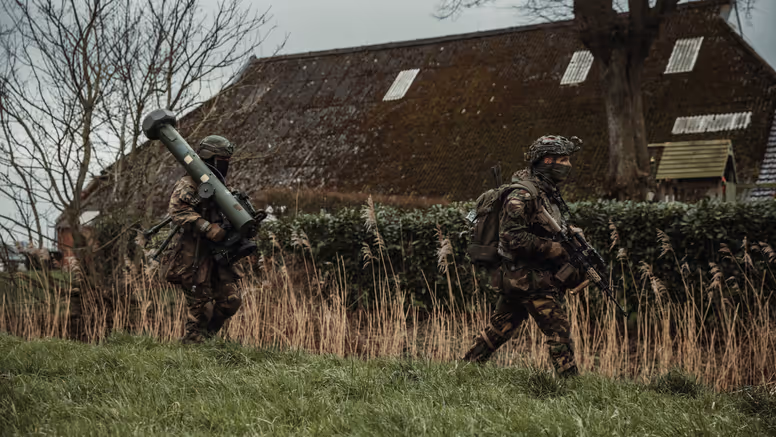 Twee gewapende militairen lopen achter elkaar door een grasveld. De achterste draagt een cilindervormig, donkergroen antitankwapen op zijn schouder.