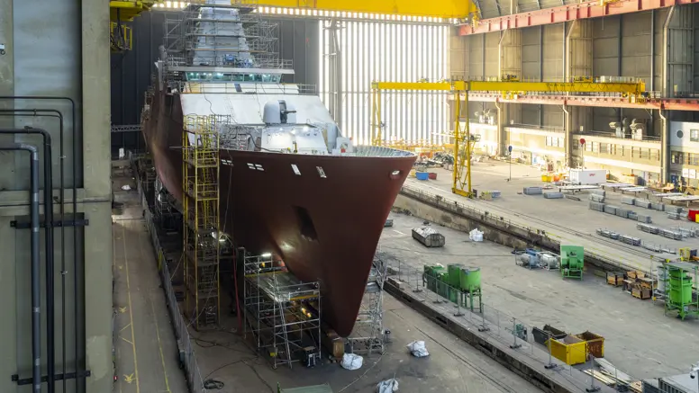 Zr.Ms. Holland in aanbouw in een overdekte scheepswerf, omringd door steigers, kranen en bouwmateriaal. 