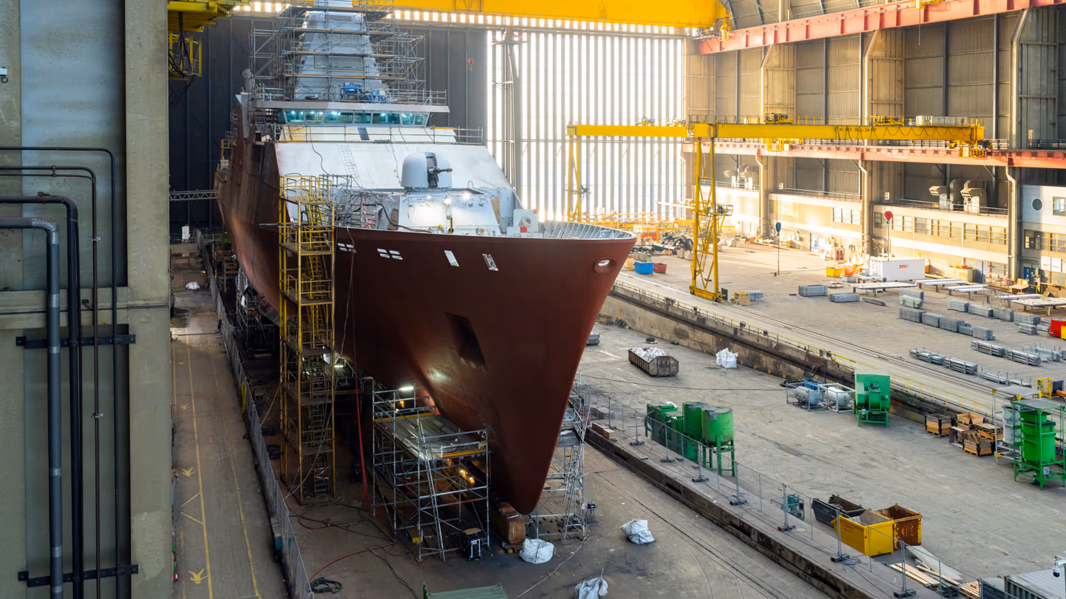 Zr.Ms. Holland in aanbouw in een overdekte scheepswerf, omringd door steigers, kranen en bouwmateriaal. 