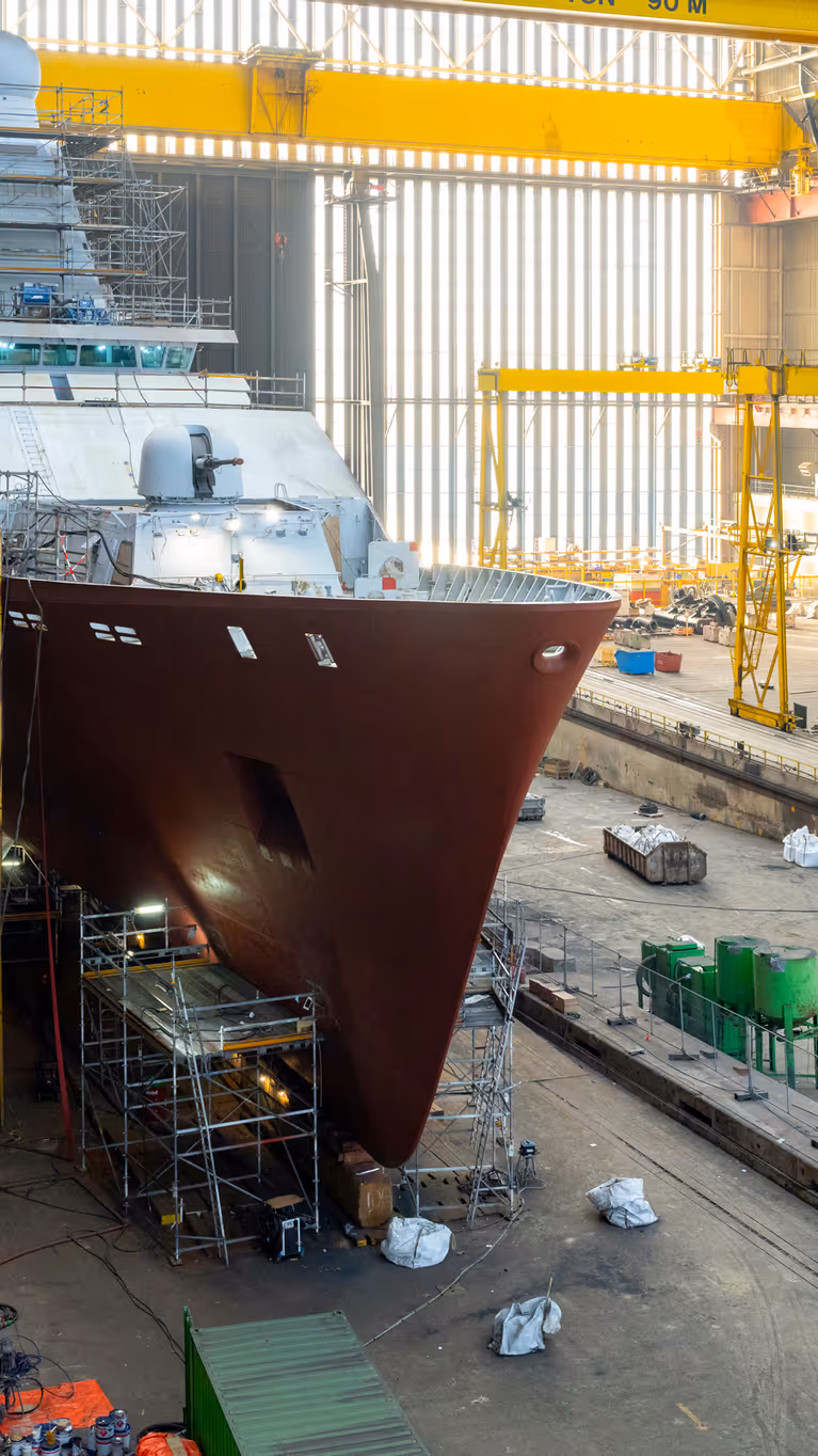 Zr.Ms. Holland in aanbouw in een overdekte scheepswerf, omringd door steigers, kranen en bouwmateriaal. 