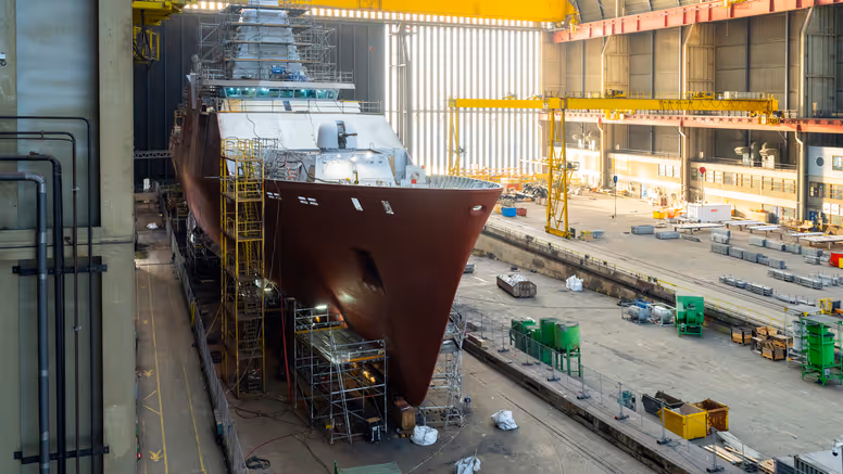 Zr.Ms. Holland in aanbouw in een overdekte scheepswerf, omringd door steigers, kranen en bouwmateriaal. 