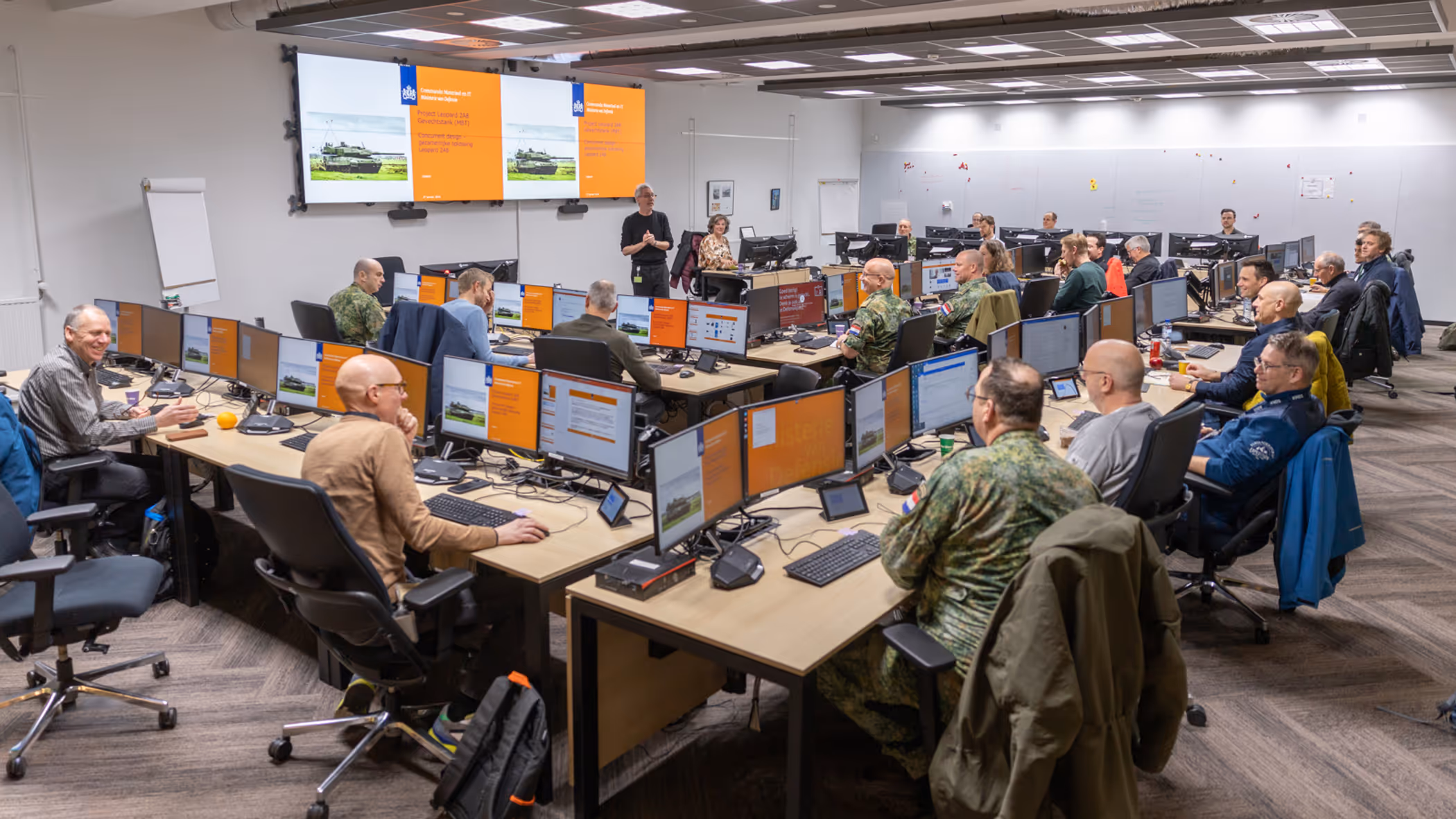 Een ruimte met daarin een dubbele u-opstelling van bureaus. Daaraan zitten militairen en burgers. Zij kijken naar een groot scherm vooraan in de ruimte, waarop een Leopard-gevechtstank te zien is.