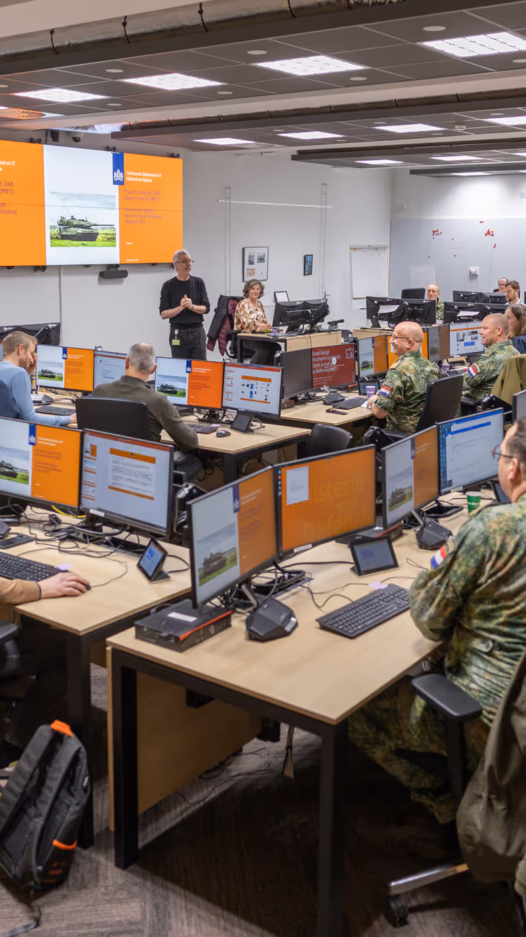Een ruimte met daarin een dubbele u-opstelling van bureaus. Daaraan zitten militairen en burgers. Zij kijken naar een groot scherm vooraan in de ruimte, waarop een Leopard-gevechtstank te zien is.