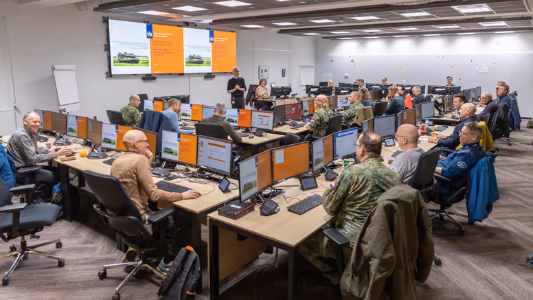 Een ruimte met daarin een dubbele u-opstelling van bureaus. Daaraan zitten militairen en burgers. Zij kijken naar een groot scherm vooraan in de ruimte, waarop een Leopard-gevechtstank te zien is.
