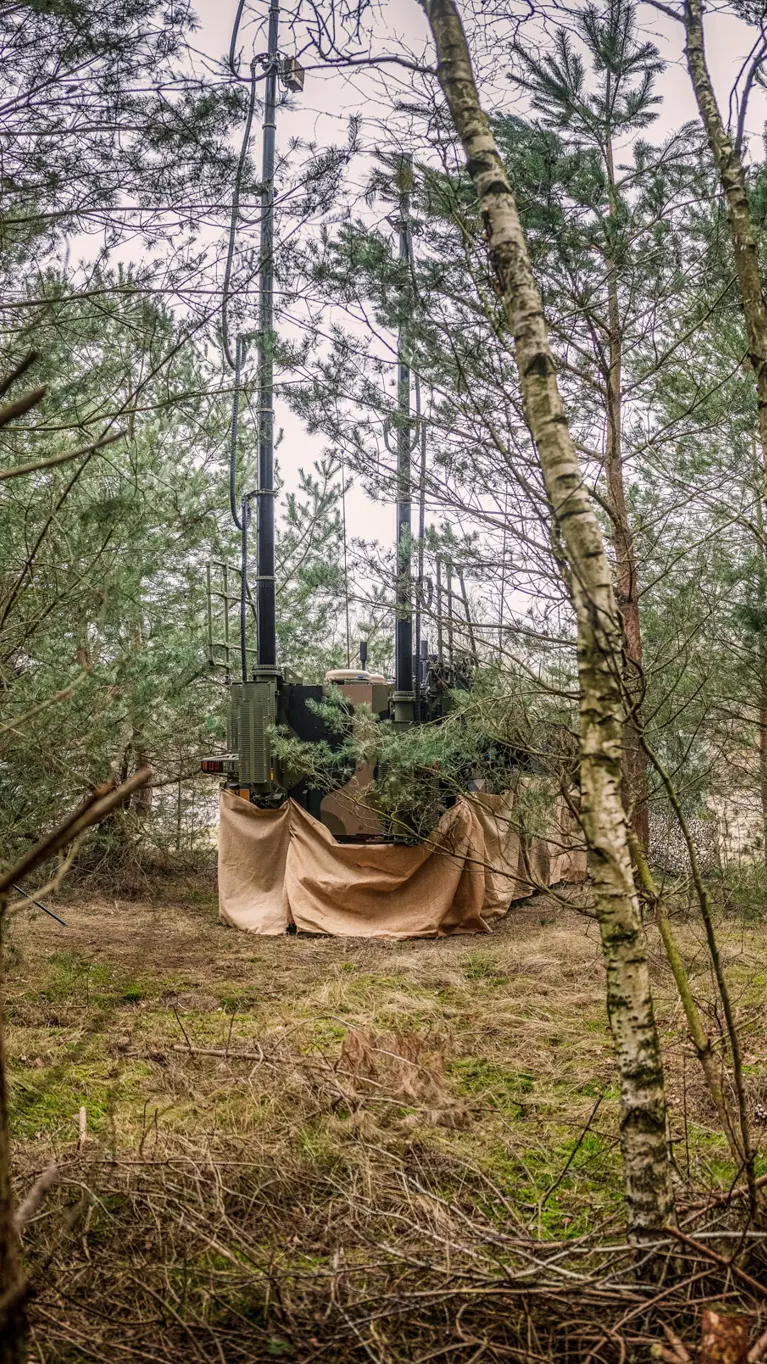 Een bewapende militair in een bosrijke omgeving. Achter hem staat een gecamoufleerd voertuig voor Elektromagnetische Oorlogvoering. Aan de onderkant is een bruin doek bevestigd.