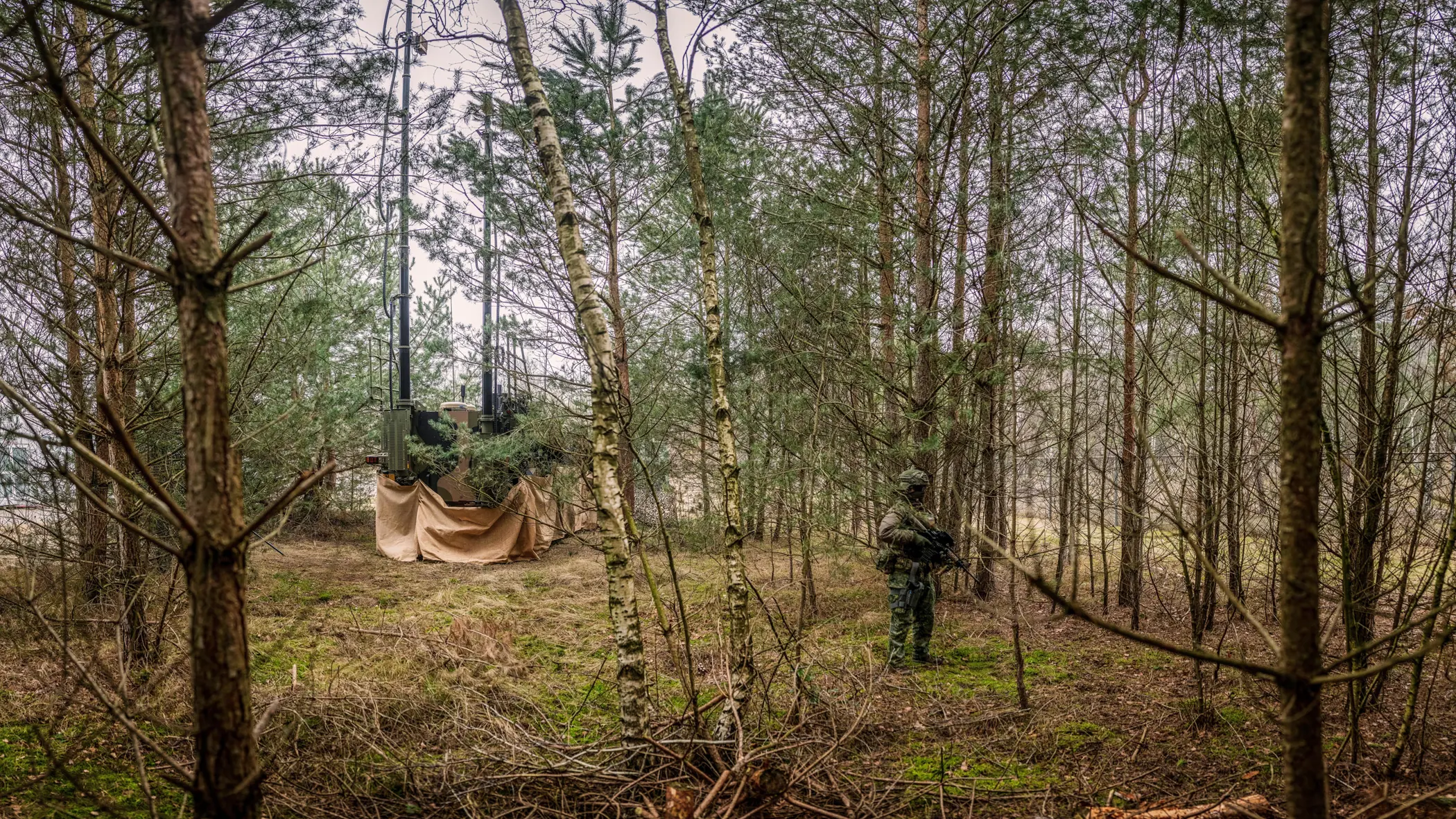 Een bewapende militair in een bosrijke omgeving. Achter hem staat een gecamoufleerd voertuig voor Elektromagnetische Oorlogvoering. Aan de onderkant is een bruin doek bevestigd.