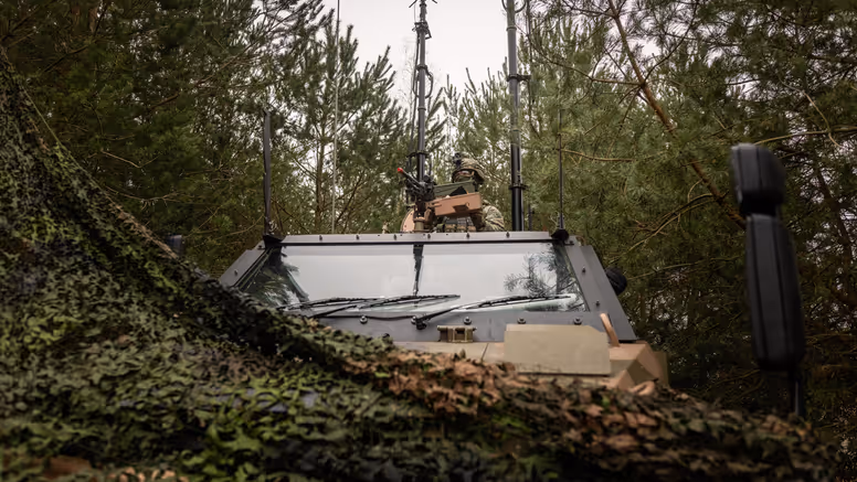Een militair zit met zijn  halve lichaam bovenop de Bushmaster. Het voertuig is bedekt met een camouflagenet en staat in bosschage. Achter de militair zijn diverse antennes te zien.