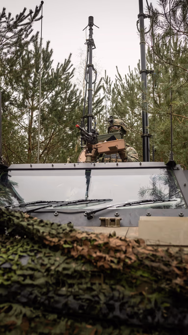 Een militair zit met zijn  halve lichaam bovenop de Bushmaster. Het voertuig is bedekt met een camouflagenet en staat in bosschage. Achter de militair zijn diverse antennes te zien.