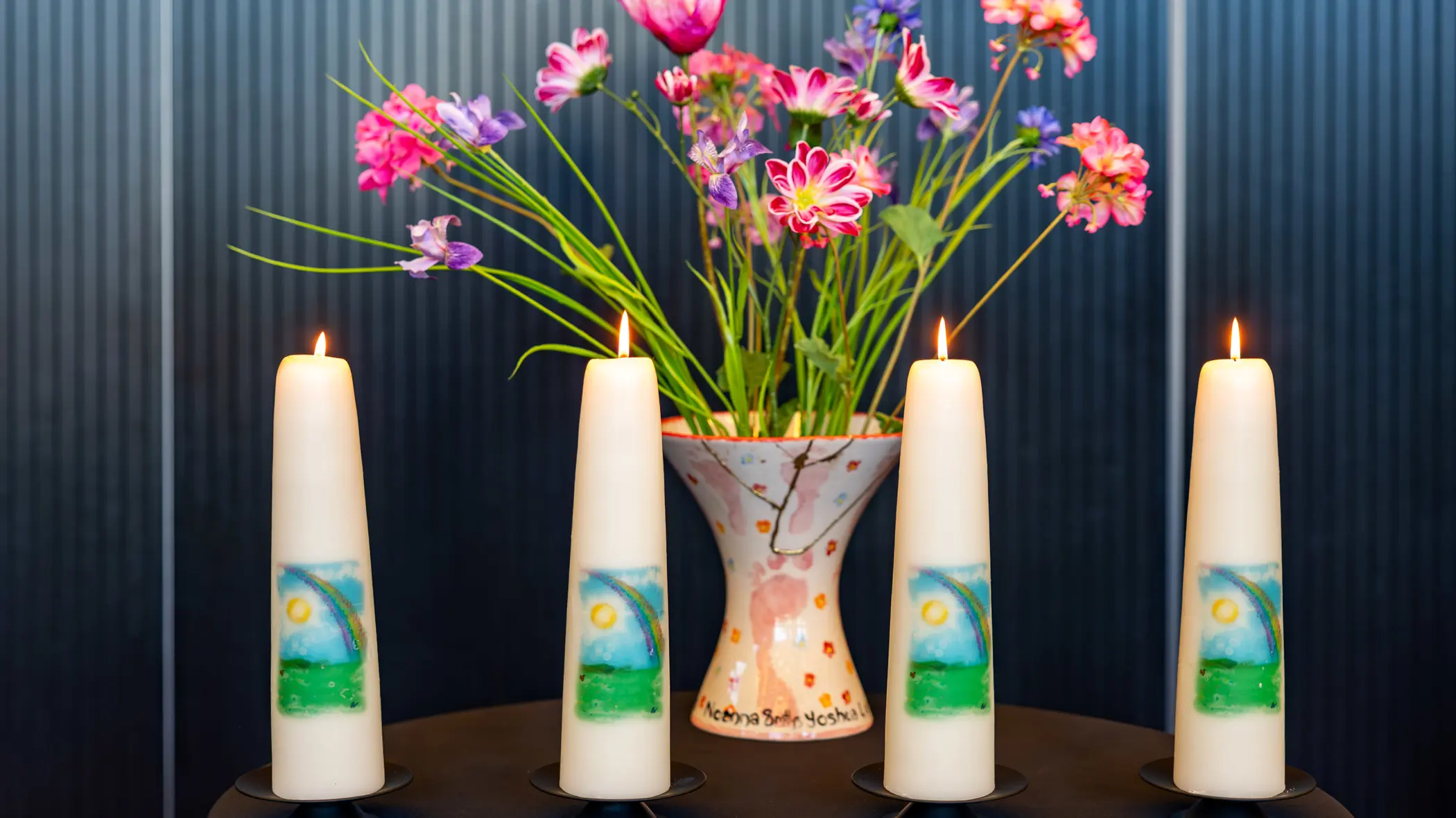 Vier brandende kaarsen op een tafel. Daarachter een vaas met gekleurde bloemen. 