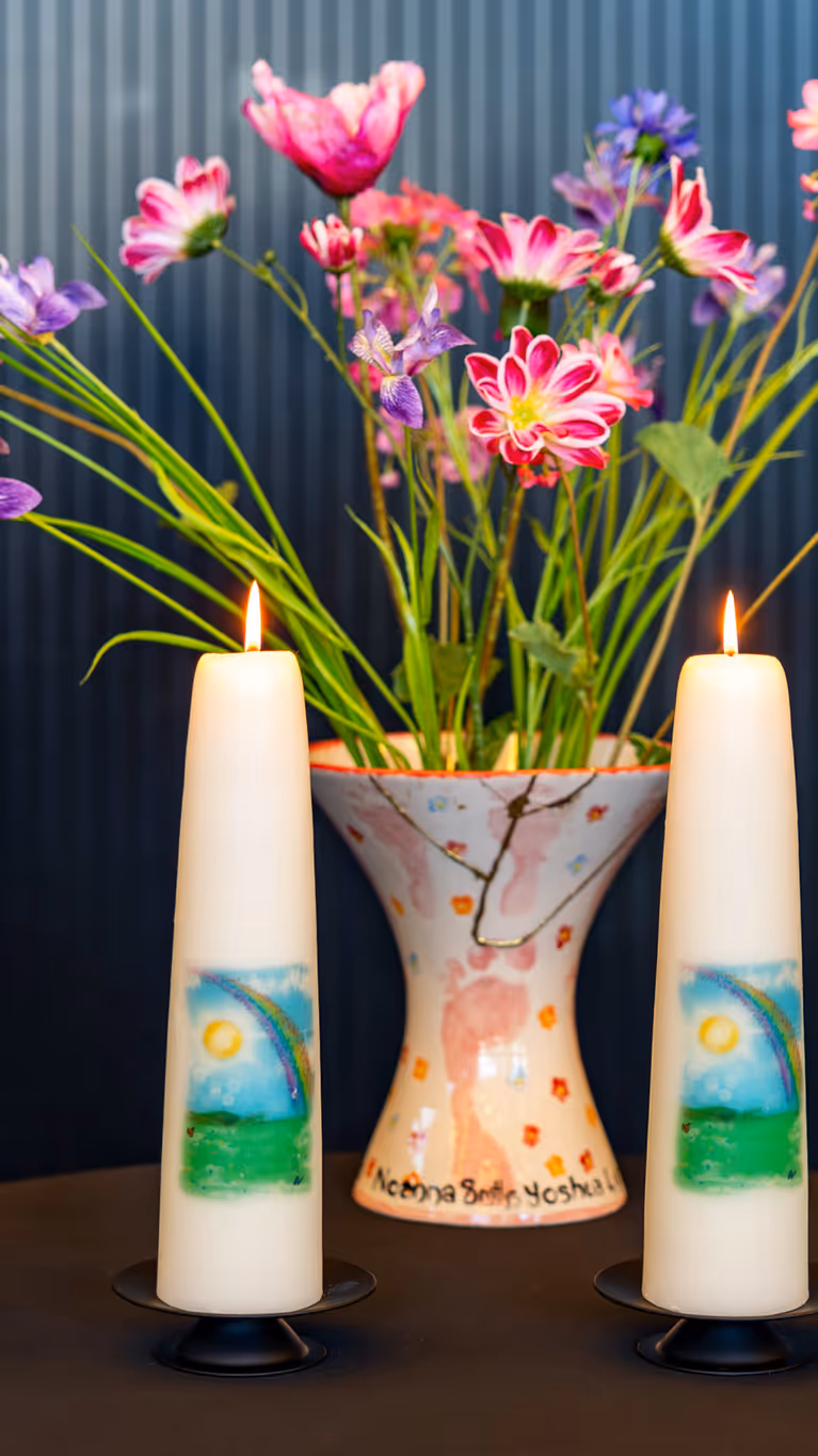 Vier brandende kaarsen op een tafel. Daarachter een vaas met gekleurde bloemen. 
