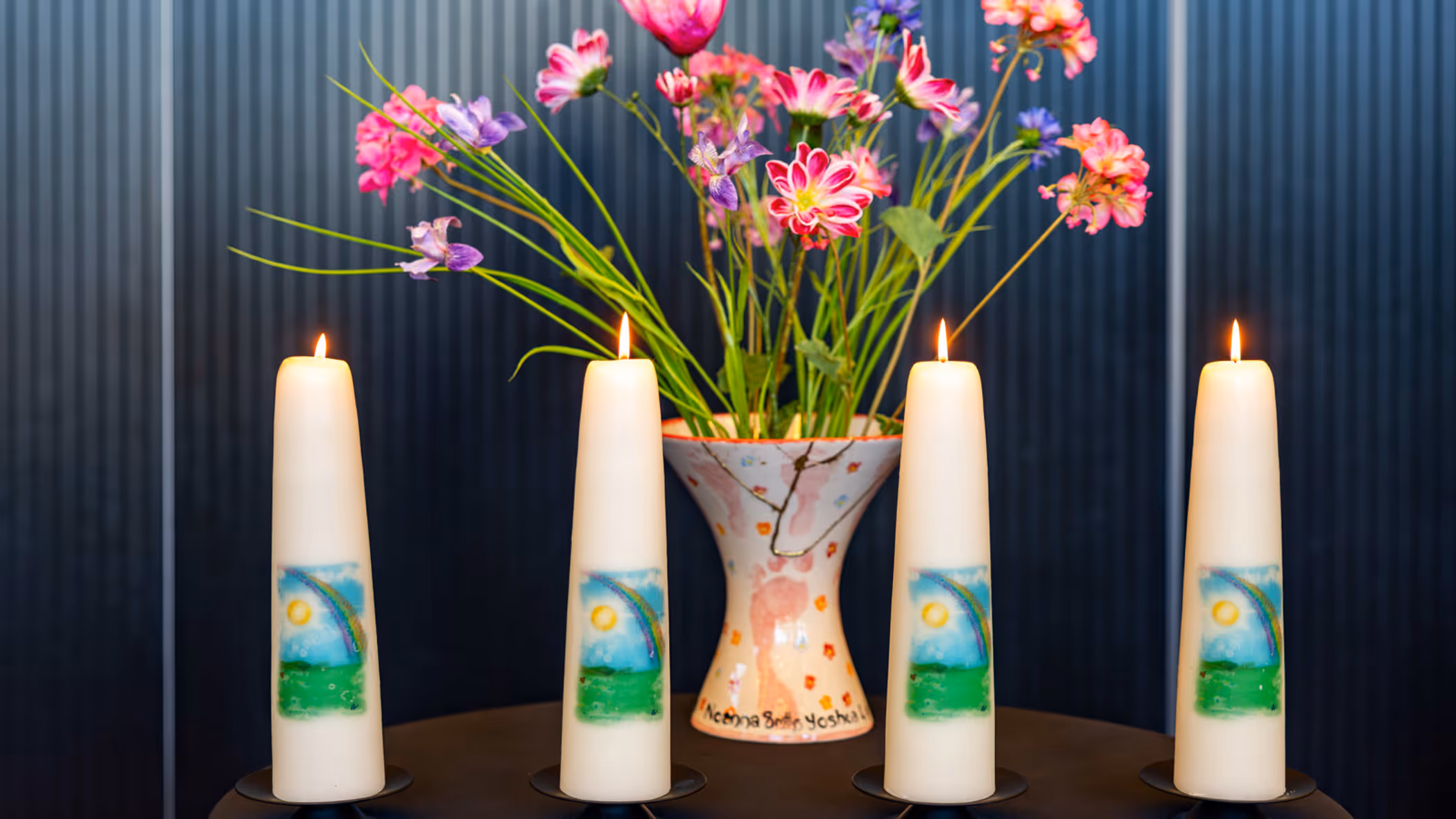 Vier brandende kaarsen op een tafel. Daarachter een vaas met gekleurde bloemen. 