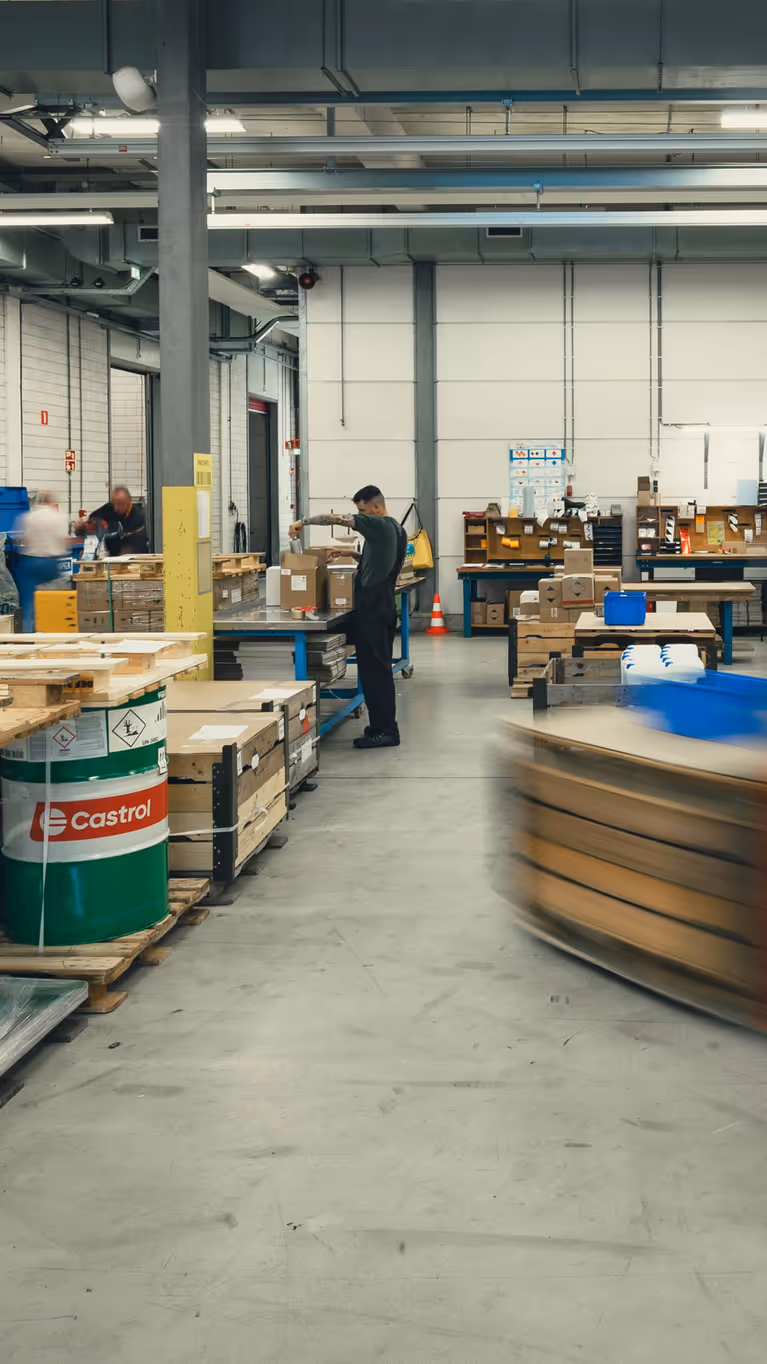 Het grijze depot van het DBBB. Hierop zijn onder meer medewerkers, pallets, vaten olie en een langsrijdende heftruck te zien.&nbsp;