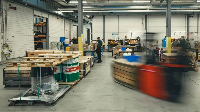 Het grijze depot van het DBBB. Hierop zijn onder meer medewerkers, pallets, vaten olie en een langsrijdende heftruck te zien.&nbsp;