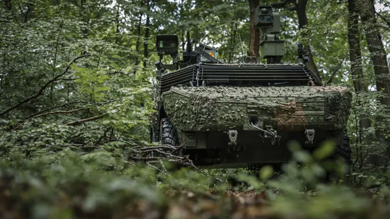 Een Fennek-voertuig rijdt door een beboste omgeving en is voorzien van een camouflagenet. 