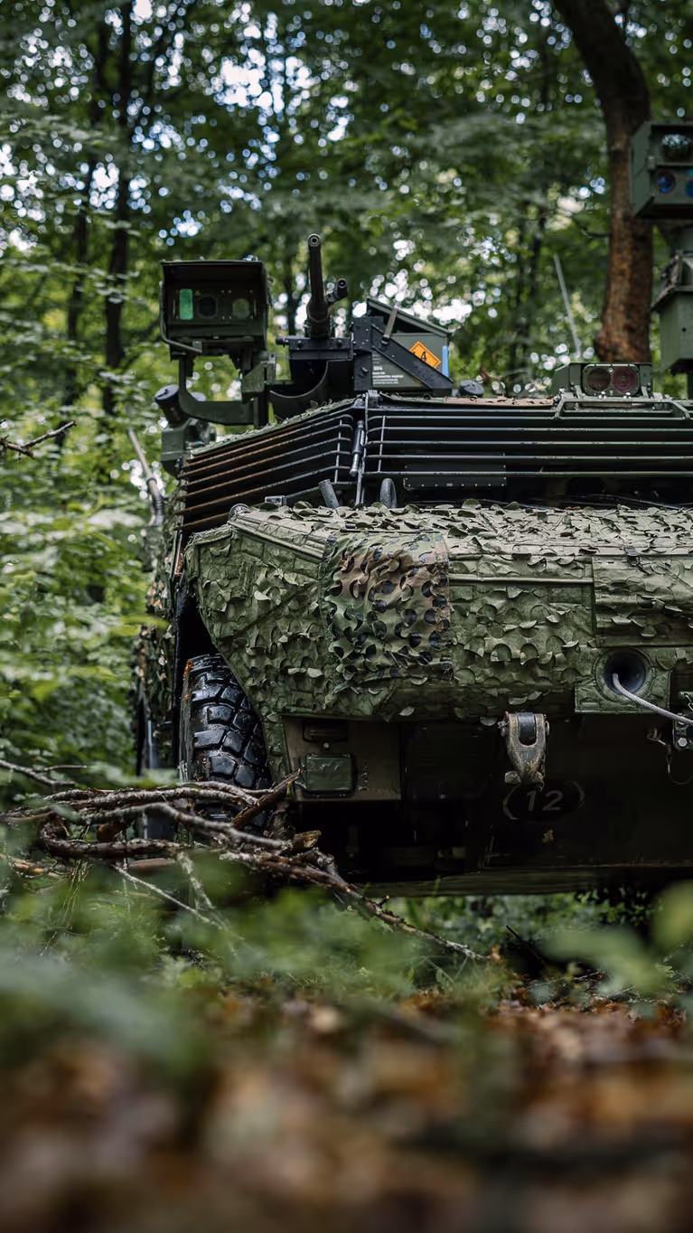 Een Fennek-voertuig rijdt door een beboste omgeving en is voorzien van een camouflagenet. 