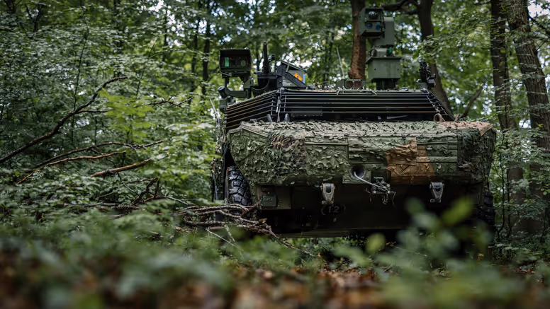 Een Fennek-voertuig rijdt door een beboste omgeving en is voorzien van een camouflagenet. 