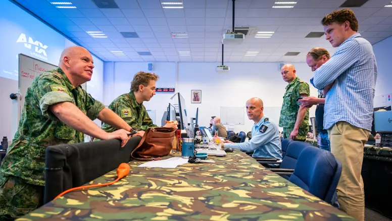 Vier militairen en twee burgermedewerkers overleggen staand en zittend aan een gecamoufleerde, bedekte tafel.