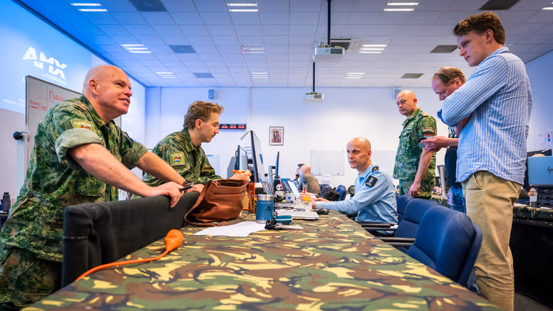 Vier militairen en twee burgermedewerkers overleggen staand en zittend aan een gecamoufleerde, bedekte tafel.