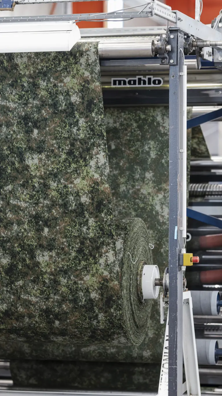Stof met groen camouflagepatroon wordt door machine geleid tijdens een controle op productiefouten.