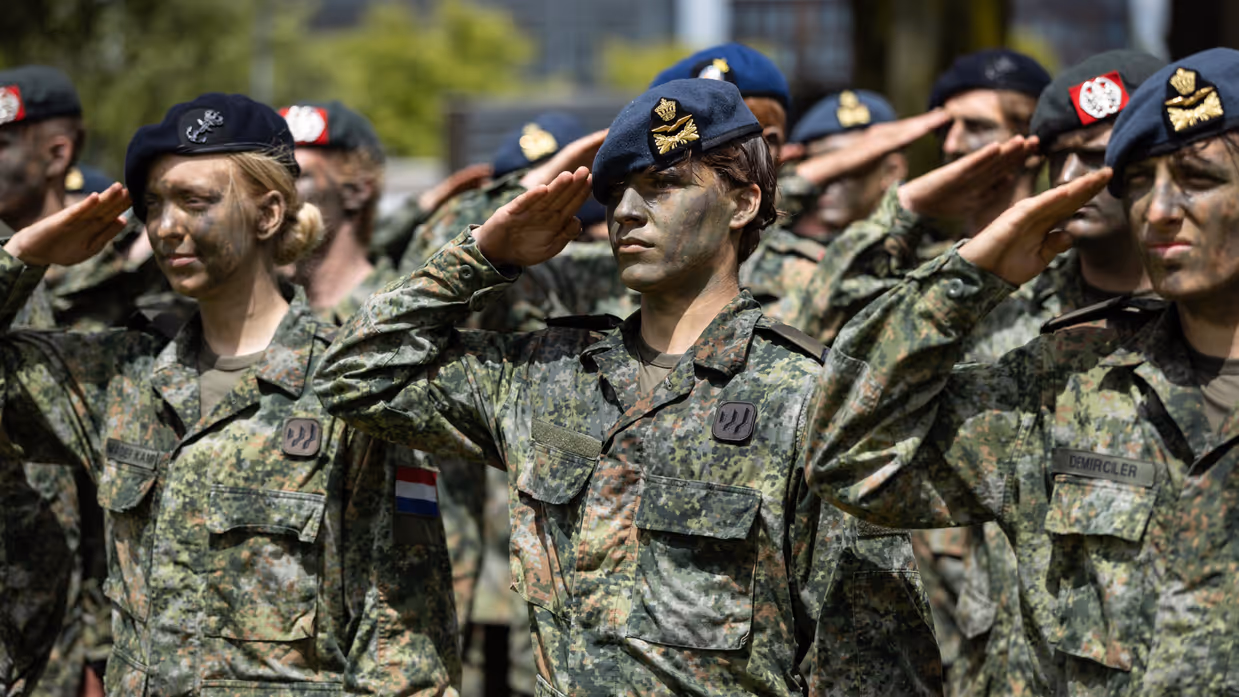 Twee luchtmachtmilitairen brengen samen met militairen van landmacht, marine en marechaussee de militaire groet. Allen met gecamoufleerde gezichten en een baret op. 