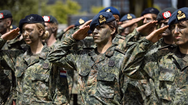 Twee luchtmachtmilitairen brengen samen met militairen van landmacht, marine en marechaussee de militaire groet. Allen met gecamoufleerde gezichten en een baret op. 