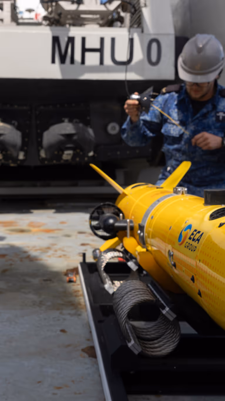 Twee marinecollega’s in blauw pak zijn op een scheepsdek bezig met een gele zeedrone. 