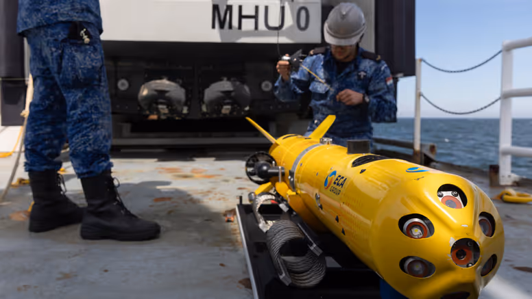 Twee marinecollega’s in blauw pak zijn op een scheepsdek bezig met een gele zeedrone. 