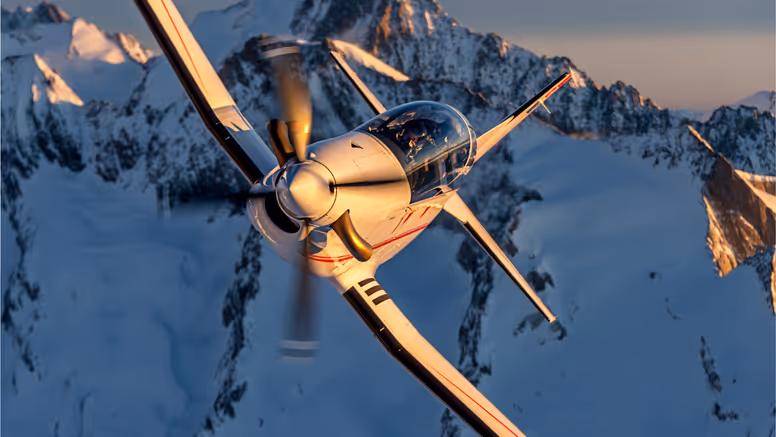 Een lesvliegtuig vliegt schuin langs winterachtige bergtoppen. 