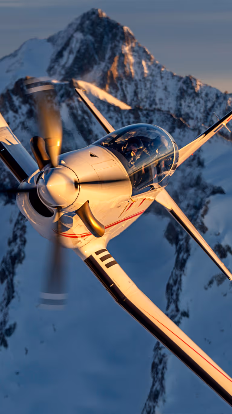 Een lesvliegtuig vliegt schuin langs winterachtige bergtoppen. 
