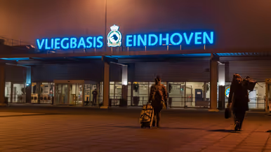 De ingang van Vliegbasis Eindhoven, af te lezen op de grote blauwe neonletters boven het gebouw. Daarnaast een vliegende C-390M.
