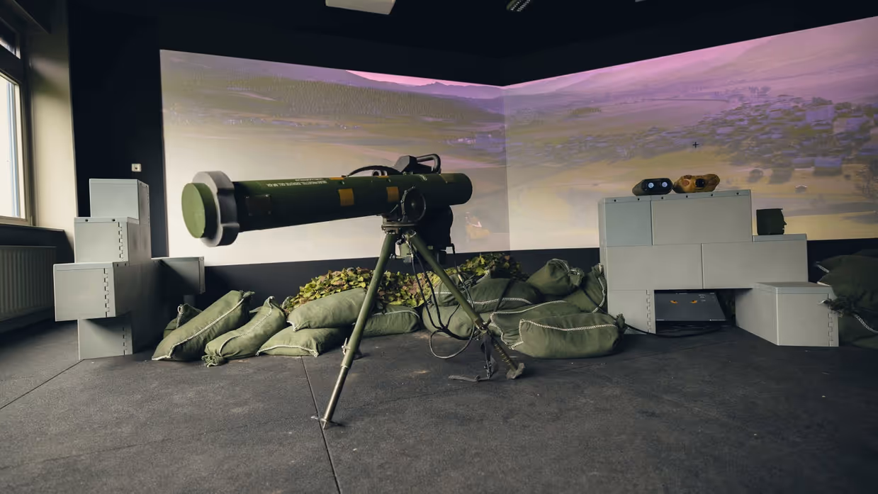 Een viertal afbeeldingen. Naast het cilindervormige, donkergroene antitankwapen Spike Long Range gericht op een beamer, hetzelfde middel alleen nu met daarachter een militair. Samen met een instructeur, die achter een beeldscherm zit, bevinden de twee zich in een kleinere ruimte. Vervolgens diezelfde oefenende militair schuin vanaf onder en gezien vanaf voren. Daarbij is zijn geconcentreerde blik zichtbaar.