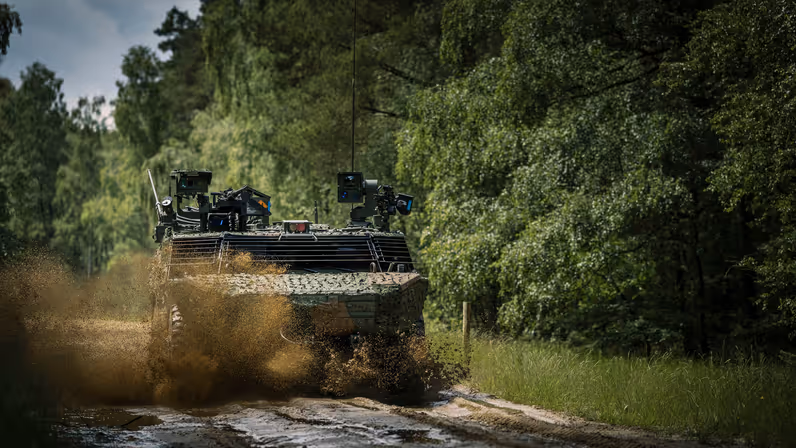Een militair voertuig raast in het bos over een zandweg. 