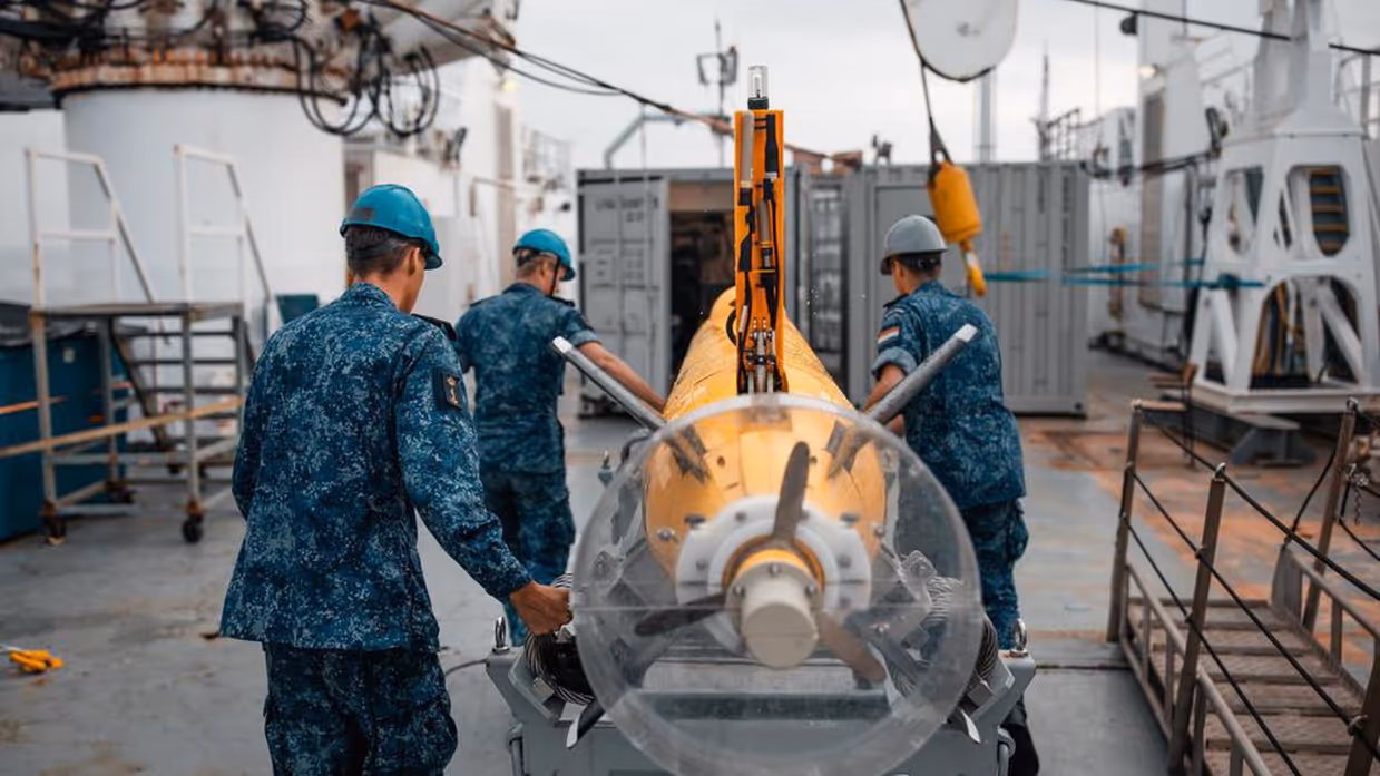 Marinecollega’s met een bouwvakkershelm op zijn bezig met een zeedrone die in de takels hangt. Op de andere foto ligt de gele zeedrone in het water, op de achtergrond zijn marineschepen te zien. 