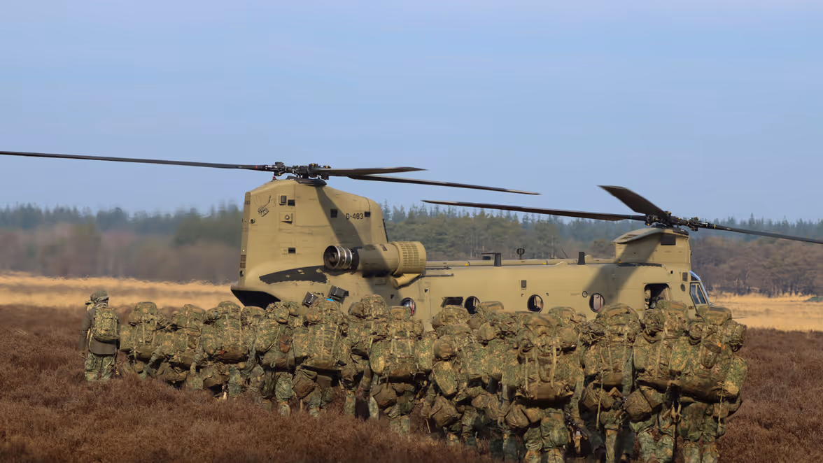 Een grote groep militairen met rugzakken en wapens lopen naar de achterzijde van een grote helikopter op de heide.